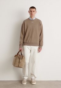 Béžový sweatshirt s rebrovanými manžetami, svetlomodrá košeľa s golierom pod ním, biele nohavice, béžové tenisky a béžová taška s tmavými rúčkami.