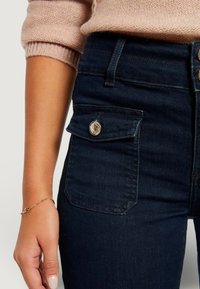 Vaqueros de mezclilla azul oscuro con un bolsillo delantero que presenta un botón decorativo plateado. Textura suave, diseño de talle alto, detalles de costura visibles.