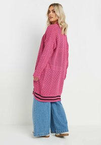 Rosa stickad cardigan med geometriska mönster, öppen front, långa ärmar och svart randig fåll. Matchad med vida blå jeans.