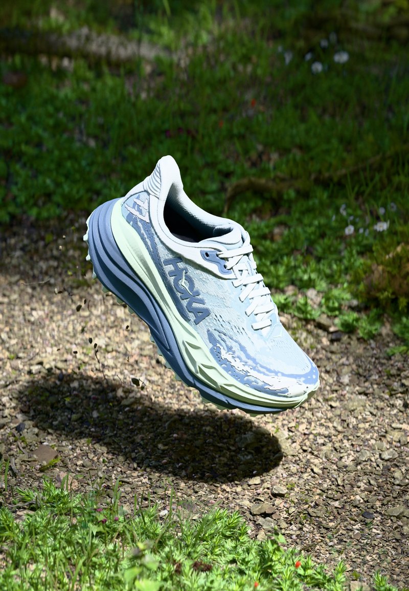 Scarpa da corsa azzurro chiaro con tomaia texturizzata, intersuola verdognola e lacci bianchi. Design elevato per un'adeguata ammortizzazione, ambientata in un contesto naturale all'aperto.