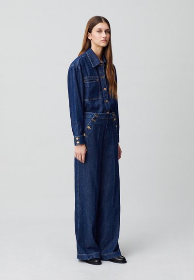 Mono de mezclilla en azul oscuro con mangas largas, parte delantera abotonada, grandes bolsillos y diseño de pernera ancha. Presenta botones dorados y una silueta estructurada.