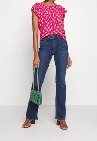 Rote Bluse mit floralem Muster und Rüschensäumen, kombiniert mit dunkelblauen Schlagjeans. Grünes Handtasche mit Krokodilprägung und braune Sandalen mit Riemchen.