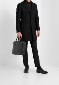 Zwarte wollen overjas met notch-lapels, knoopsluiting en zijzakken; gecombineerd met strakke broek en zwarte leren handtas voor een stijlvolle look.