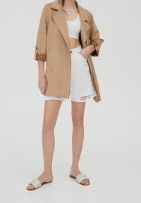 Femme portant un manteau beige sur un crop top blanc, un short en jean blanc déchiré, et des sandales slides beiges, debout devant un fond uni.