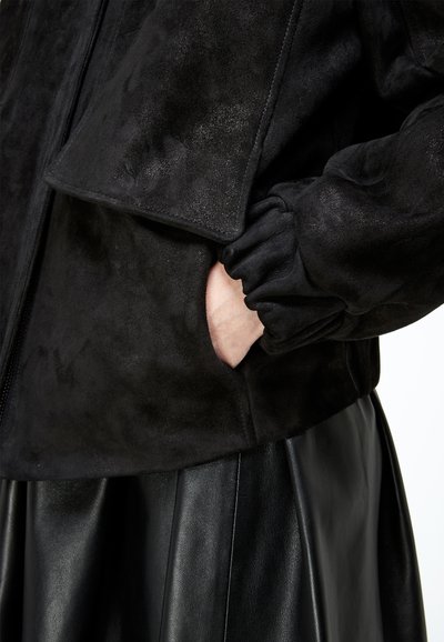 Personne portant une veste en suède noire avec un poignet élastique, main dans la poche latérale, assortie à une jupe plissée en cuir noir.
