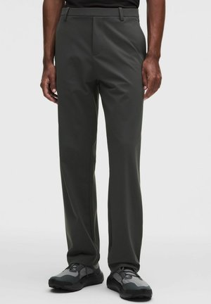 Mann trägt dunkelgraue Hose mit geradem Bein und grau-schwarze Sneakers, steht vor einfachem hellem Hintergrund.