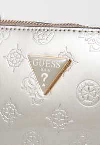 Silver bag featuring embossed patterns, a triangular logo plaque with "GUESS U.S.A.," and a textured zipper along the top.