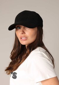 Vrouw met een zwarte corduroy baseballcap en een wit T-shirt, met lang bruin haar, die lichtjes opzij kijkt tegen een effen achtergrond.