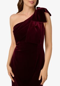 Abito in velluto bordeaux con design a spalla unica, caratterizzato da tessuto plissettato e un grande fiocco decorativo sulla spalla. Texture liscia.