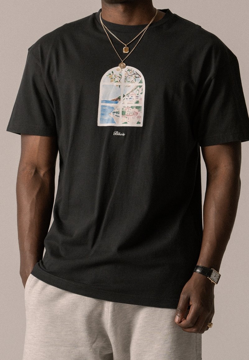 Homme portant un t-shirt noir avec une scène de fenêtre d’une ville côtière, deux colliers en or, un pantalon beige, une montre et une bague.