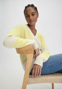Strickpullover mit gelb-beigen Streifen, V-Ausschnitt, strukturiertem Stoff und gerippten Bündchen, kombiniert mit blauen Jeans auf einem Holstuhl.