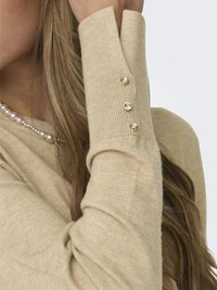 Pull en tricot beige à manches longues, doté de trois boutons décoratifs sur le poignet. Texture douce et design ajusté.