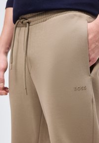Μπεζ παντελόνια φόρμας BOSS με κορδόνι στη μέση, πλαϊνή τσέπη με το χέρι μέσα, φορεμένα με σκούρο τοπ.