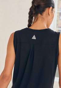 Schwarzes ärmelloses Oberteil mit einem plissierten Design auf der Rückseite und einem weißen Dreieck-Logo. Glatte Textur mit einem lässigen Schnitt.
