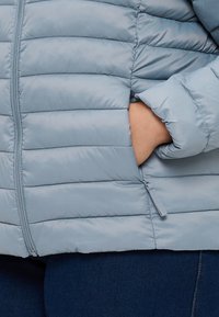 Gaiši zila pūkainā jaka ar horizontālu quilted dizainu, ar rāvējslēdzēju priekšā un sānu kabatām. Gluda tekstūra. Valkāta virs tumšām džinsa biksēm.