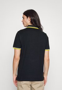 Camisa polo preta com colarinho e punhos em amarelo riscado, com mangas curtas e um corte solto, feita de um tecido texturado e suave.
