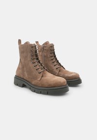 Bottines en suede marron à lacets sur le devant, avec une fermeture éclair latérale et des semelles épaisses en caoutchouc noir. Présente des coutures renforcées et une languette à l'arrière.