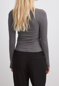 Femme aux longs cheveux blonds portant un haut gris côtelé à manches longues ajusté et un pantalon noir, vue de dos sur un fond uni.
