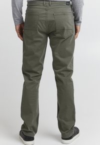 Olijfgroene chinos met een rechte pijp, voorzien van twee achterzakken en een taillebandlabel, gecombineerd met zwarte sneakers. Gladde textuur.