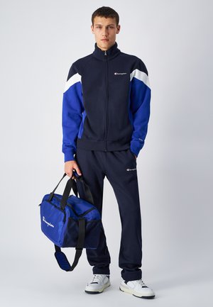 Chándal navy con acentos en azul y blanco, que incluye una chaqueta con cremallera y pantalones a juego. Una bolsa de deporte azul es sostenida por la modelo en la mano.