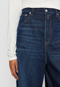 Person som har på seg mørkeblå denimjeans og en hvit langermet skjorte, med hånden avslappet ved siden av.