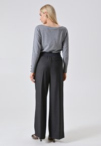 Donna con lunghi capelli biondi, che indossa un top grigio a maniche lunghe, pantaloni larghi a vita alta scuri e scarpe con tacco, in piedi di spalle.