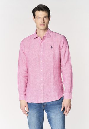 Uomo che indossa una camicia rosa a maniche lunghe con piccolo logo sul petto e jeans blu, in piedi davanti a uno sfondo bianco semplice.
