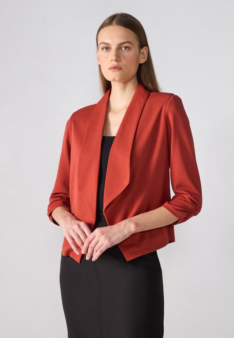 Rust-colored cropped blazer with a draped front, three-quarter sleeves, and ruched details. Worn over a black dress. Smooth fabric texture.