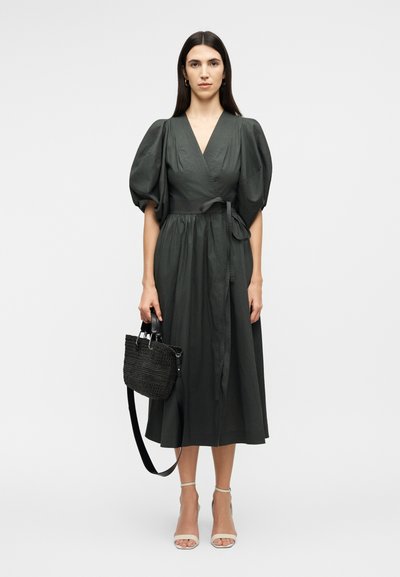 Γυναίκα με σκούρο midi φόρεμα τύπου wrap με φουσκωτά μανίκια, κρατώντας μια μαύρη τσάντα με υφή και φορόντας μπεζ πέδιλα με λουράκι στον αστράγαλο.