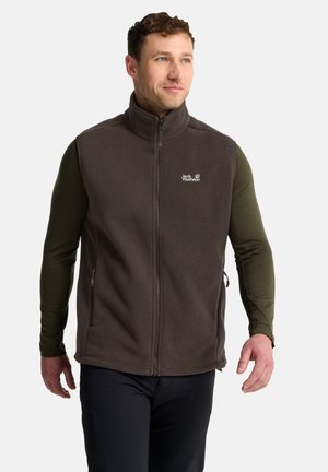 Mann mit lockigem Haar trägt eine braune Fleeceweste mit Reißverschluss über einem grünen Langarmshirt und steht vor weißem Hintergrund.