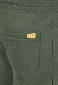 Green cotton joggers featuring a ribbed waistband, a back pocket, and a visible yellow label with the text “!SOLID.” Soft texture.