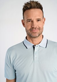 Ljusblå pikétröja med en strukturerad tygkvalitet, med en vit krage med marinblå detaljer och tre knappar vid halsringningen.