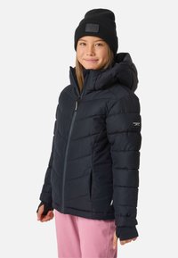 Giacca puffer blu navy con cappuccio, caratterizzata da cuciture orizzontali e polsini elasticizzati. Indossata con pantaloni rosa e un berretto nero.