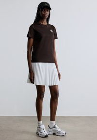 Brūna kokvilnas T-krekls ar nelielu grafiku, kopā ar baltu plisušu svārku. Valkāts ar baltām sporta kurpēm un melnu cepuri.