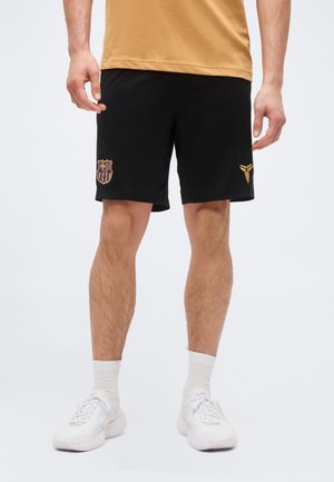 Homme portant un short de sport noir avec les logos dorés du FC Barcelone et de Kobe Bryant, des chaussettes blanches et des chaussures de sport blanches, debout devant un fond uni.
