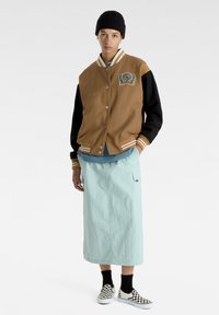 Brun og sort varsity-jakke med stribede manchetter, mintgrøn nederdel, skakbræts slip-ons, båret med en sort beanie.