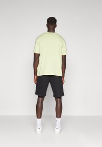 Ljust gult kortärmat t-shirt med avslappnad passform, kombinerat med svarta shorts. Modellen står mot en enkel ljus bakgrund.