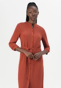 Robe chemise couleur rouille avec col montant, manches longues et une ceinture assortie. Fabriquée dans un tissu lisse avec une coupe légèrement décontractée.