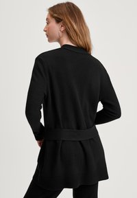 Vrouw met lang haar, gekleed in een zwarte cardigan met riem, die zijwaarts tegen een effen witte achtergrond staat.