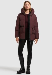 Chaqueta acolchada burdeos con cuello alto, cierre de cremallera, cintura con cordón elástico y detalles en negro. Combinada con leggings negros y botas hasta la rodilla.