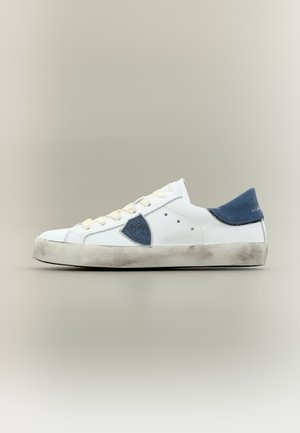 Sneaker basse blanche avec talon et empiècement latéraux bleus, lacets écrus et semelle usée et sale sur fond neutre.