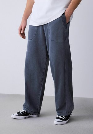 Graue Weitbein-Hose aus weichem Stoff mit zwei vorderen Taschen. Gestylt mit schwarzen und weißen Low-Top-Sneakern.