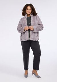 Jasnofioletowa kurtka ze sztucznego futra z wysokim kołnierzem, prążkowaną fakturą i kieszeniami na zamek, zestawiona z szarą bluzką w groszki i ciemnymi spodniami.