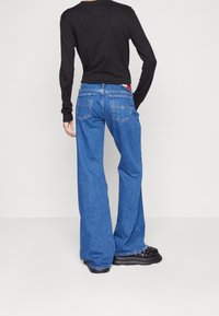 Blue denim wide-leg jeans with a mid-rise, featuring two back pockets and a branded waistband. Paired with a black long-sleeve top.