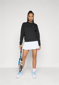 Svart hettegenser, hvit tenniskjørt med perforert tekstur, holder en blå og svart tennisklubb, sportslige hvite joggesko og ankelstrømper.