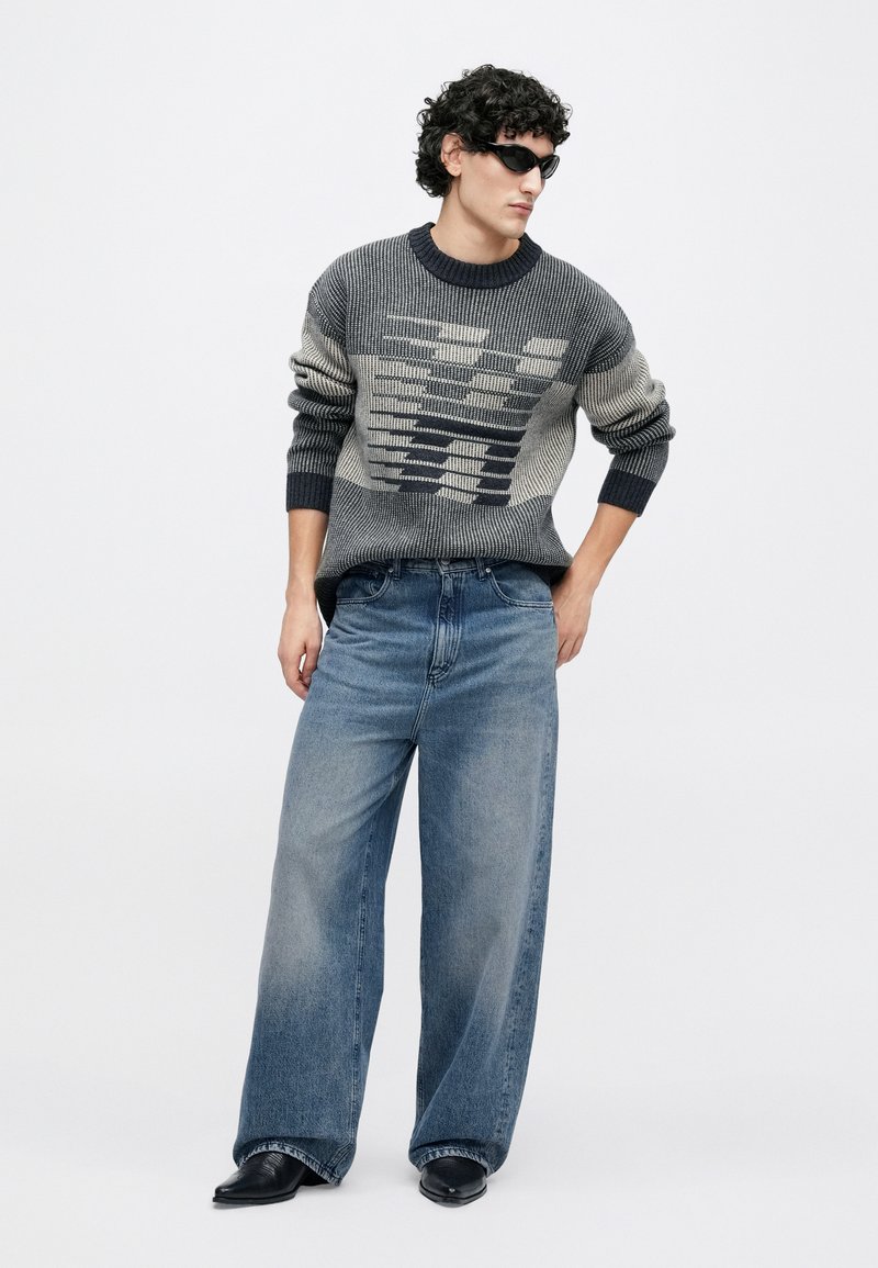 Pull en maille gris et crème avec motif géométrique, associé à un jean large bleu clair. Chaussures noires pointues et lunettes de soleil complètent le look.