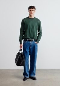 Νέος άνδρας που στέκεται με το χέρι στην τσέπη, φοράει πράσινο πουλόβερ, μπλε τζιν, μαύρα παπούτσια, κρατώντας μαύρη τσάντα ταξιδίου μπροστά σε απλό φόντο.