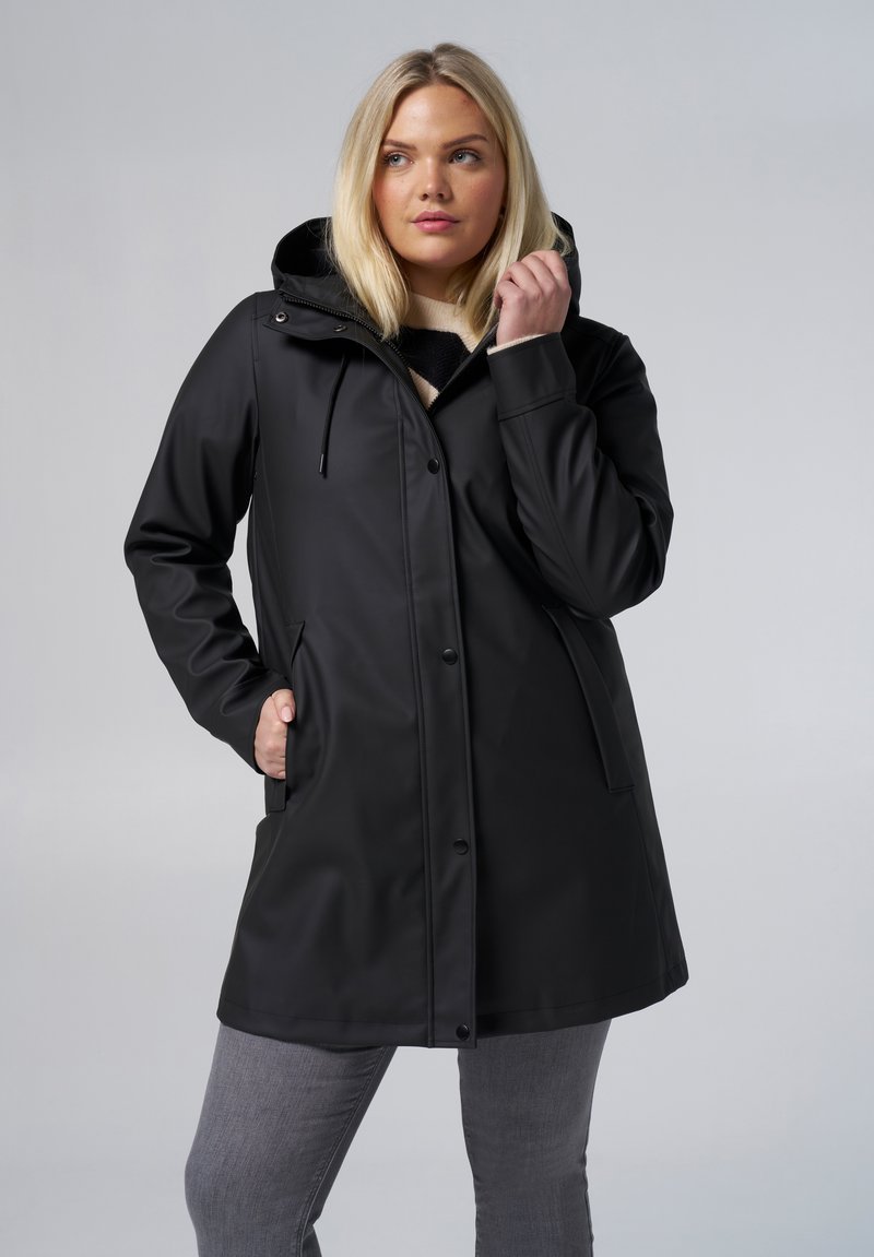 Manteau noir imperméable avec capuche, doté d'une fermeture éclair, de poches latérales et d'une texture lisse. Porté par-dessus un pull à rayures et un pantalon gris.