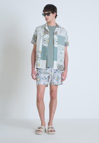 Korte mouw multi-patroon shirt in wit en groene tinten, gecombineerd met gepatternde shorts. Model draagt een zonnebril en lichte sandalen.