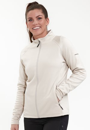 Vaaleanbeige fleece-takki, jossa on korkea kaulus, vetoketj закрыт, ja kaksi sivutaskua. Ominaisuuksiin kuuluvat litteät saumat ja tiukka istuvuus.